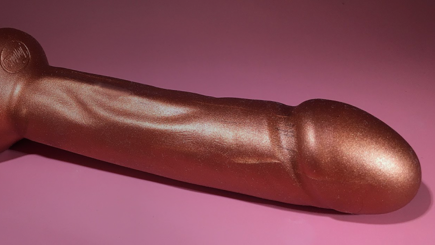 Un dildo anal gigante de color oro rosado de marca Tantus sobre fondo rosado